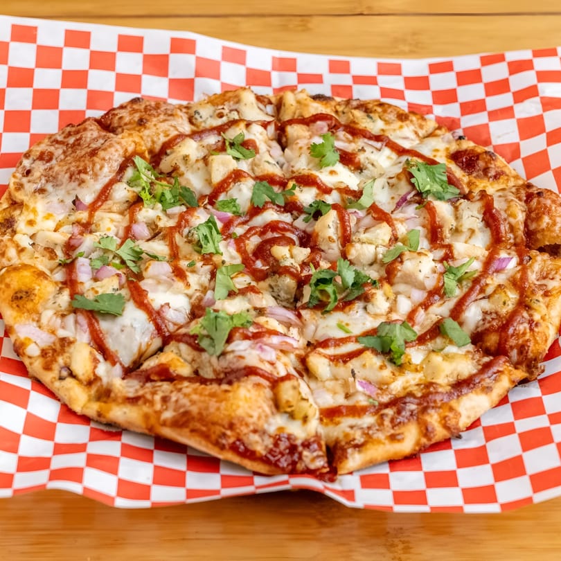 BBQ chicken pizza with red onion, cilantro, and BBQ sauce on red and white checkered paper.