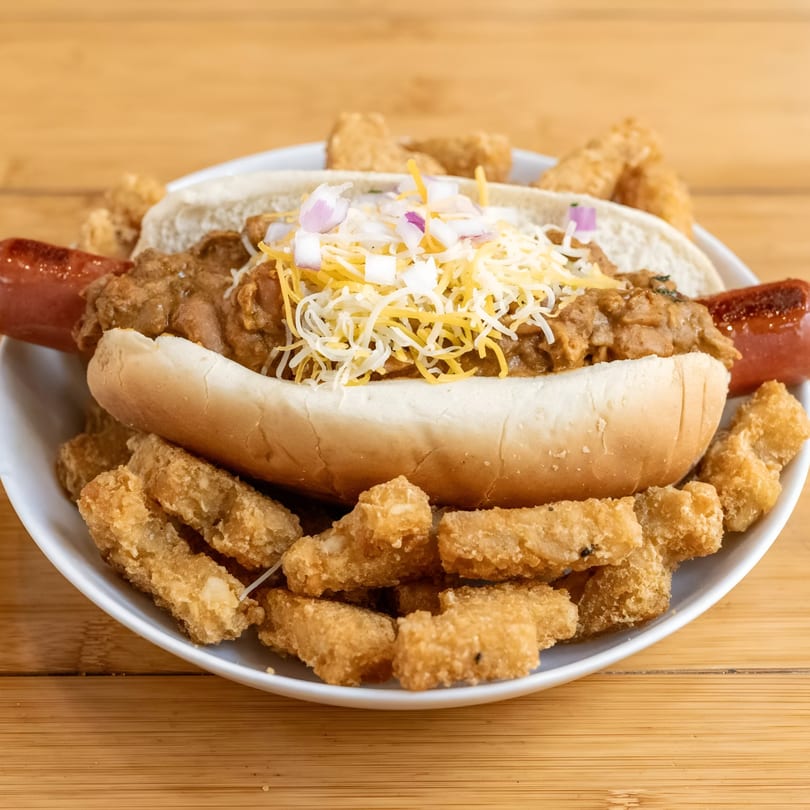 Chili dog with shredded cheese and diced onions, surrounded by tater tots on a white plate.