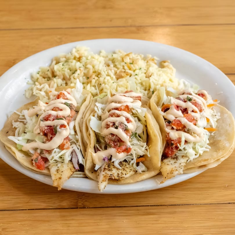 Three fish tacos topped with pico de gallo, shredded cabbage, and creamy sauce, served with rice.