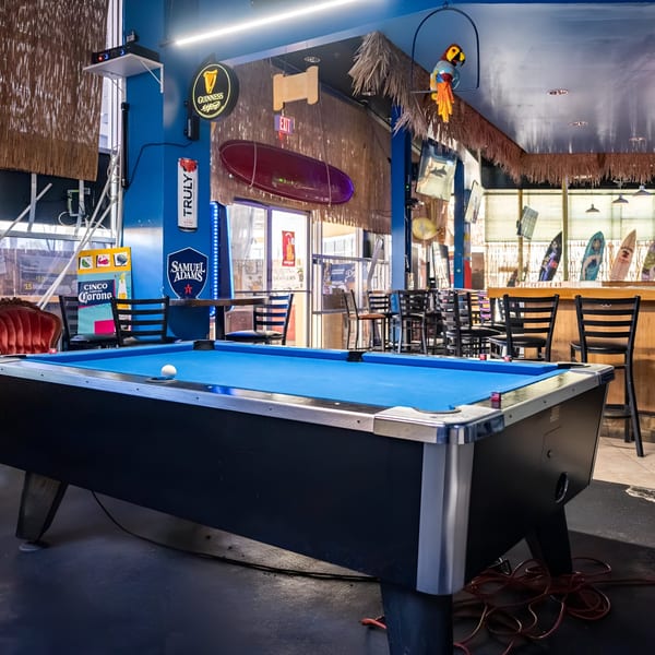 A blue-felt pool table in a bar with surfboards, bar stools, and Guinness and Samuel Adams signs.