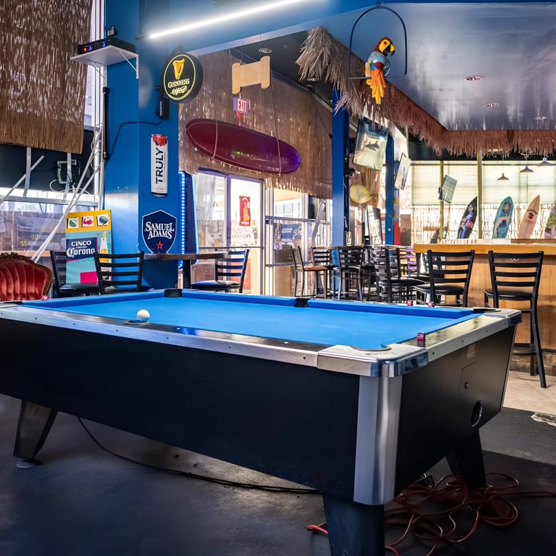 A blue-felt pool table in a bar with surfboards, bar stools, and Guinness and Samuel Adams signs.