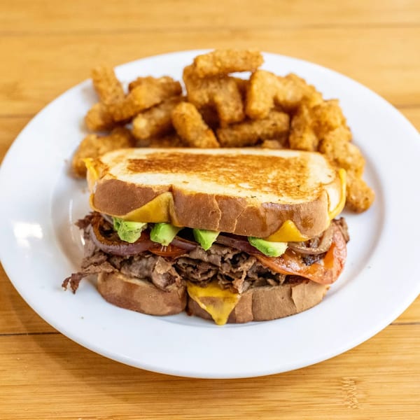 Grilled sandwich with steak, avocado, and tomatoes, served with tater tots on a white plate.